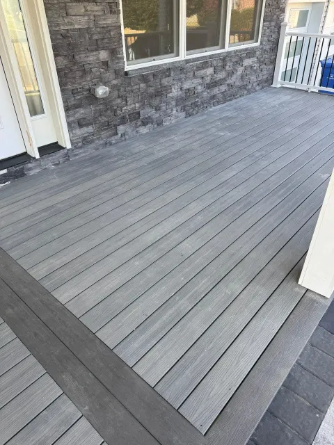 A deck with gray composite boards bordered by a darker frame, positioned against a house with stone-textured siding.