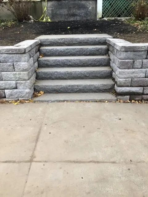 Stone steps with retaining walls leading up from a concrete sidewalk to a higher yard.
