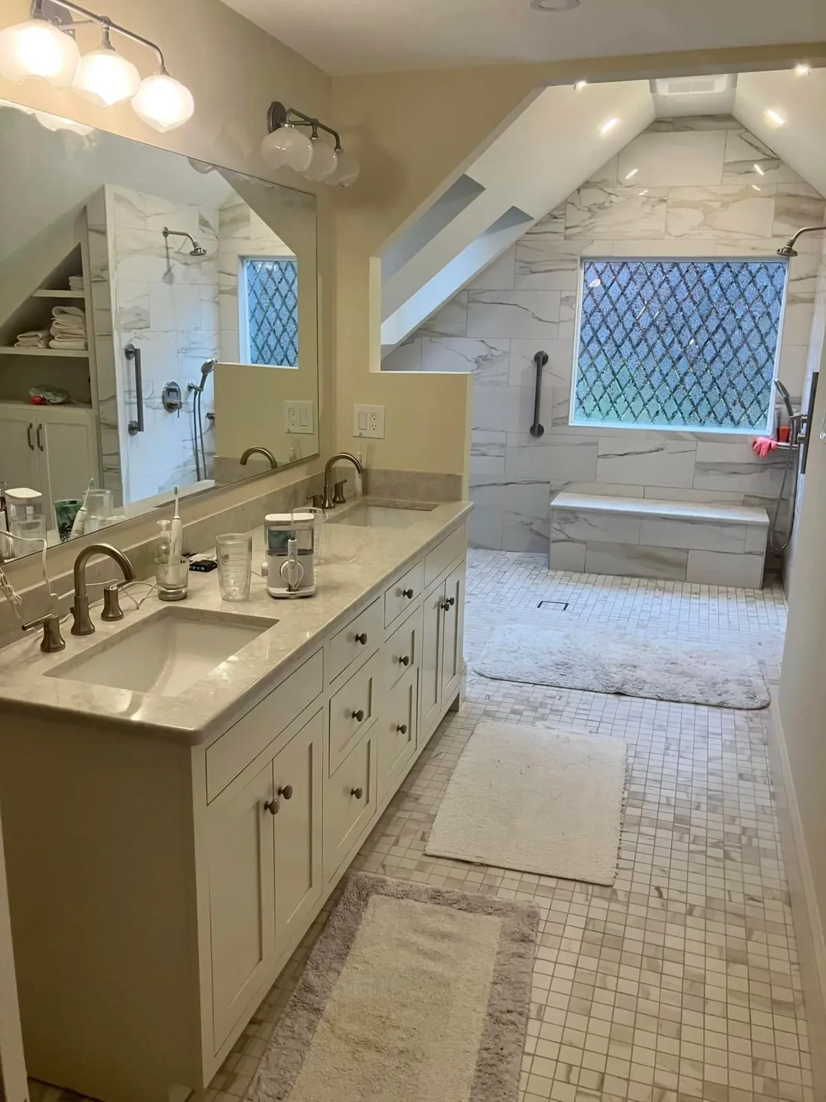 Bathroom with double vanity, large mirror, marble countertops, four bathroom rugs, walk-in shower with bench and window, and sloped ceiling with recessed lighting.