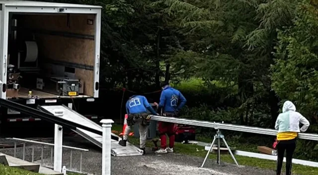 Chris Battaini Roofing - Workers load a metal beam onto a truck in a wooded area. A person in a yellow jacket stands nearby, and metal structures are visible around them.