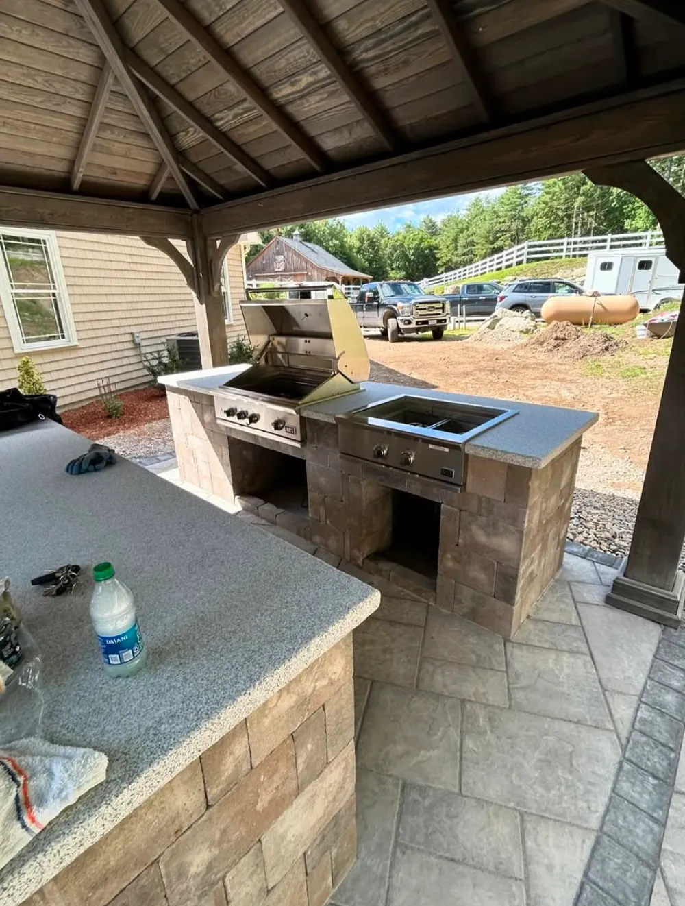 Outdoor patio area with a built-in stone barbecue grill under a wooden pergola. The countertop has a water bottle and keys on it. The setting includes a view of a yard with parked cars and trees in the background.