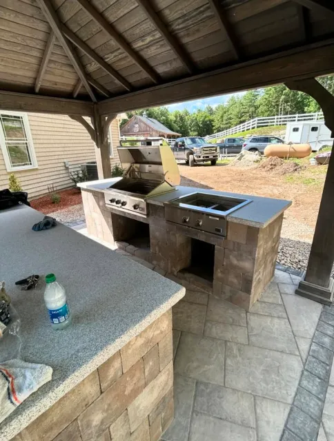 Outdoor patio area with a built-in stone barbecue grill under a wooden pergola. The countertop has a water bottle and keys on it. The setting includes a view of a yard with parked cars and trees in the background.
