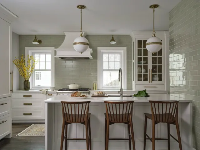 Modern kitchen with white cabinetry, green subway tile backsplash, and dark wood bar stools at a white kitchen island. Two white pendant lights hang above the island, and a window above the sink shows a snowy outdoor scene. Decor includes a vase of yellow flowers, a loaf of bread on a plate, and eggs.