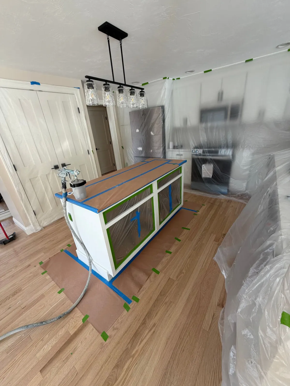 A kitchen island is taped off and being prepped for painting, with a spray gun sitting on top and plastic sheeting nearby.