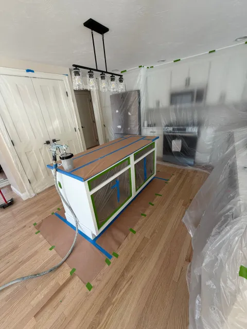 A kitchen island is taped off and being prepped for painting, with a spray gun sitting on top and plastic sheeting nearby.