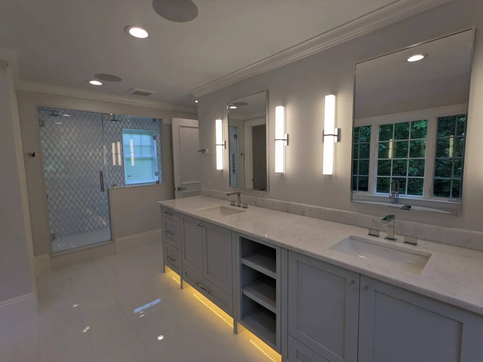 Modern bathroom with double vanity, two mirrors, vertical light fixtures, a walk-in shower with patterned glass, and a large window with lush greenery outside.