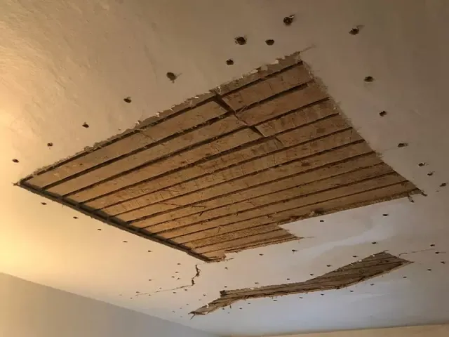 A ceiling with damaged plaster showing underlying wooden lath, with multiple holes and a larger rectangular section exposed.