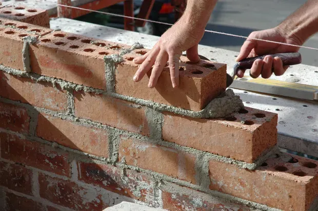 Hands laying red bricks on a partially built brick wall with mortar and tools nearby
