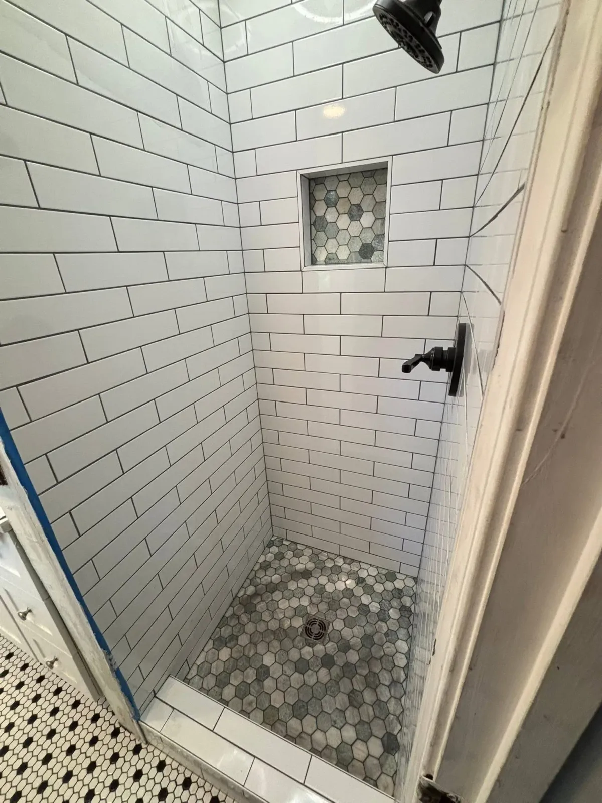 Inside a tiled shower with white subway tiles on the walls, a small hexagon tile shower floor with a drain, a built-in soap niche with hexagon tiles, and a black showerhead.