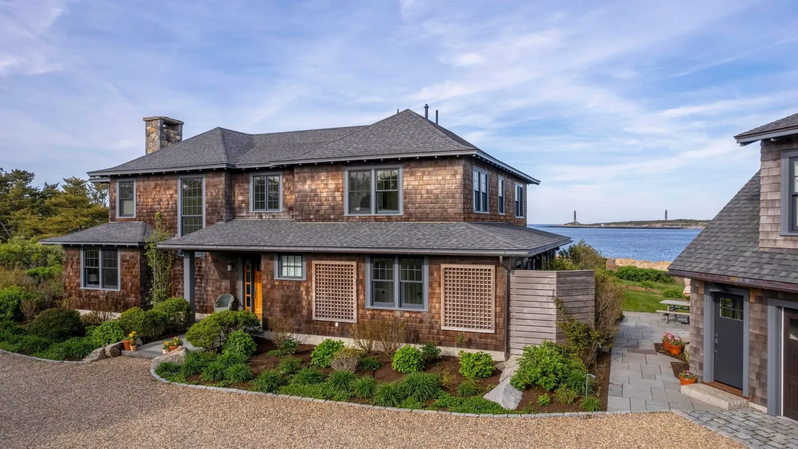 A large, shingle-style house, surrounded by a landscaped garden, overlooking a body of water with small islands and lighthouses in the distance.