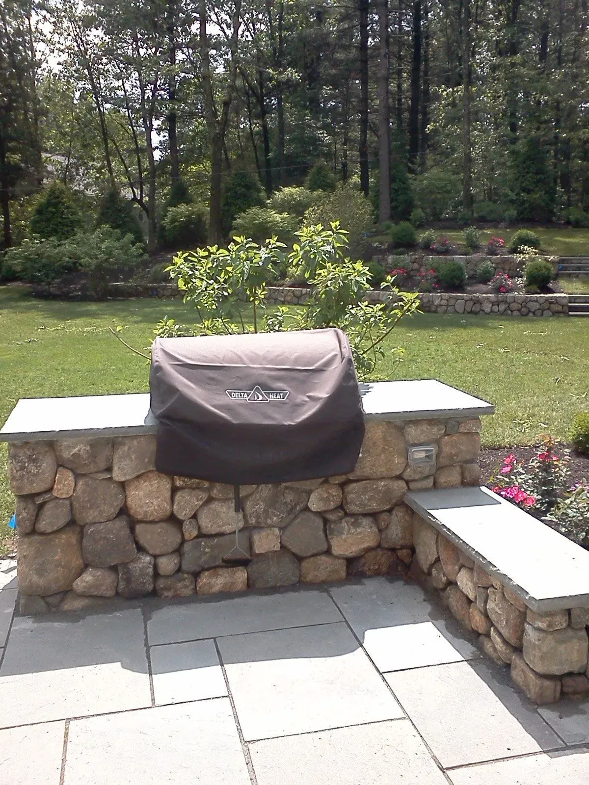 Backyard patio with stone grill wall, covered grill, lawn, flower bed, and trees.