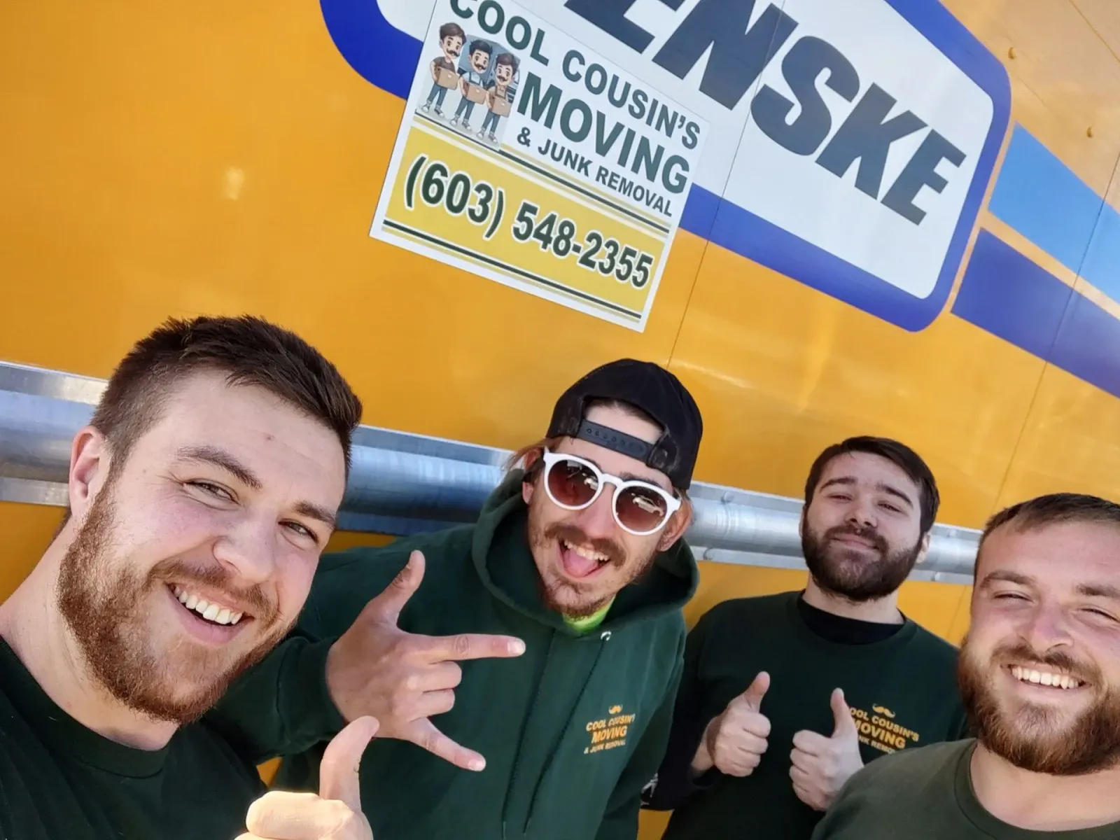 Four smiling men posing in front of a yellow Penske moving truck sign, giving thumbs-up and rock hand gestures