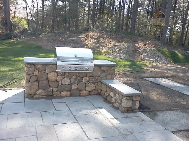 Stone outdoor grill with stainless lid and built-in bench on a paved patio near trees