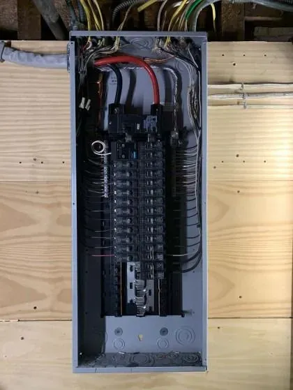 An open electrical panel on a wooden wall; black and silver circuits and wires are visible.