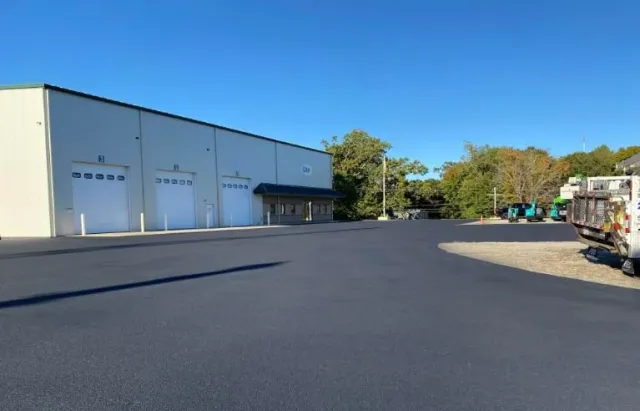 Industrial warehouse with three garage doors, a small entrance, and a freshly paved parking lot under a clear blue sky.