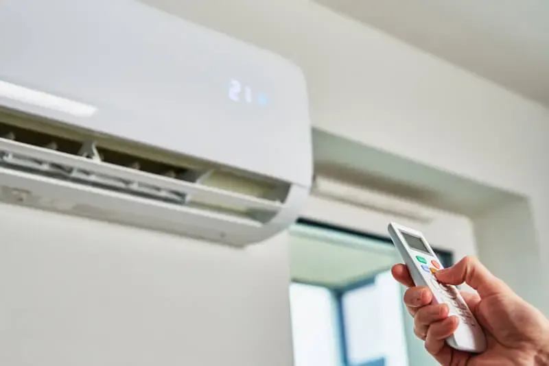 Person holds a remote control and aims it at a wall-mounted white air conditioner unit.
