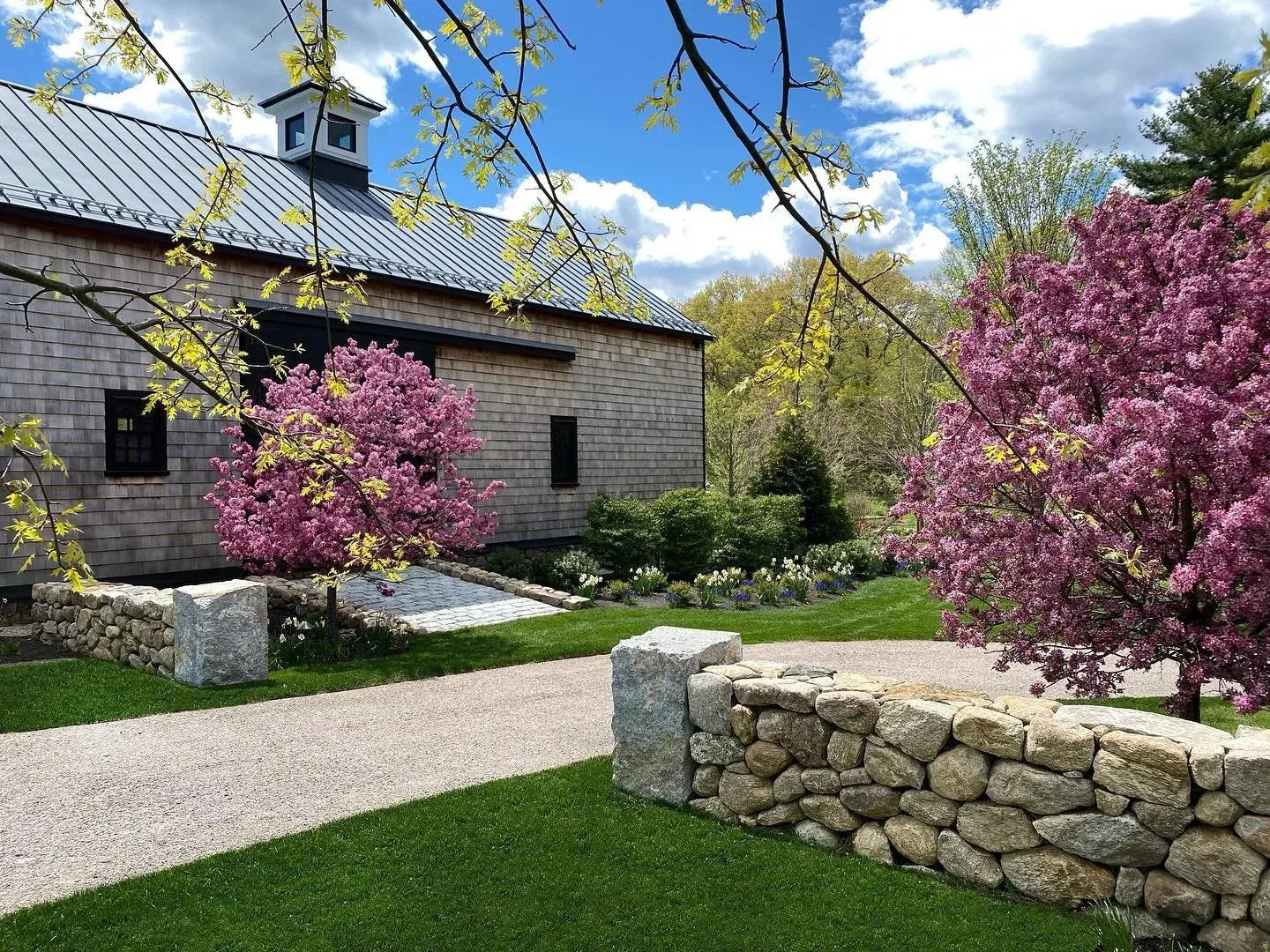 A garden scene with a stone wall, pink flowering trees, a gravel pathway, and a wooden building with a metal roof under a partly cloudy sky.