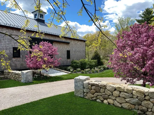 A garden scene with a stone wall, pink flowering trees, a gravel pathway, and a wooden building with a metal roof under a partly cloudy sky.