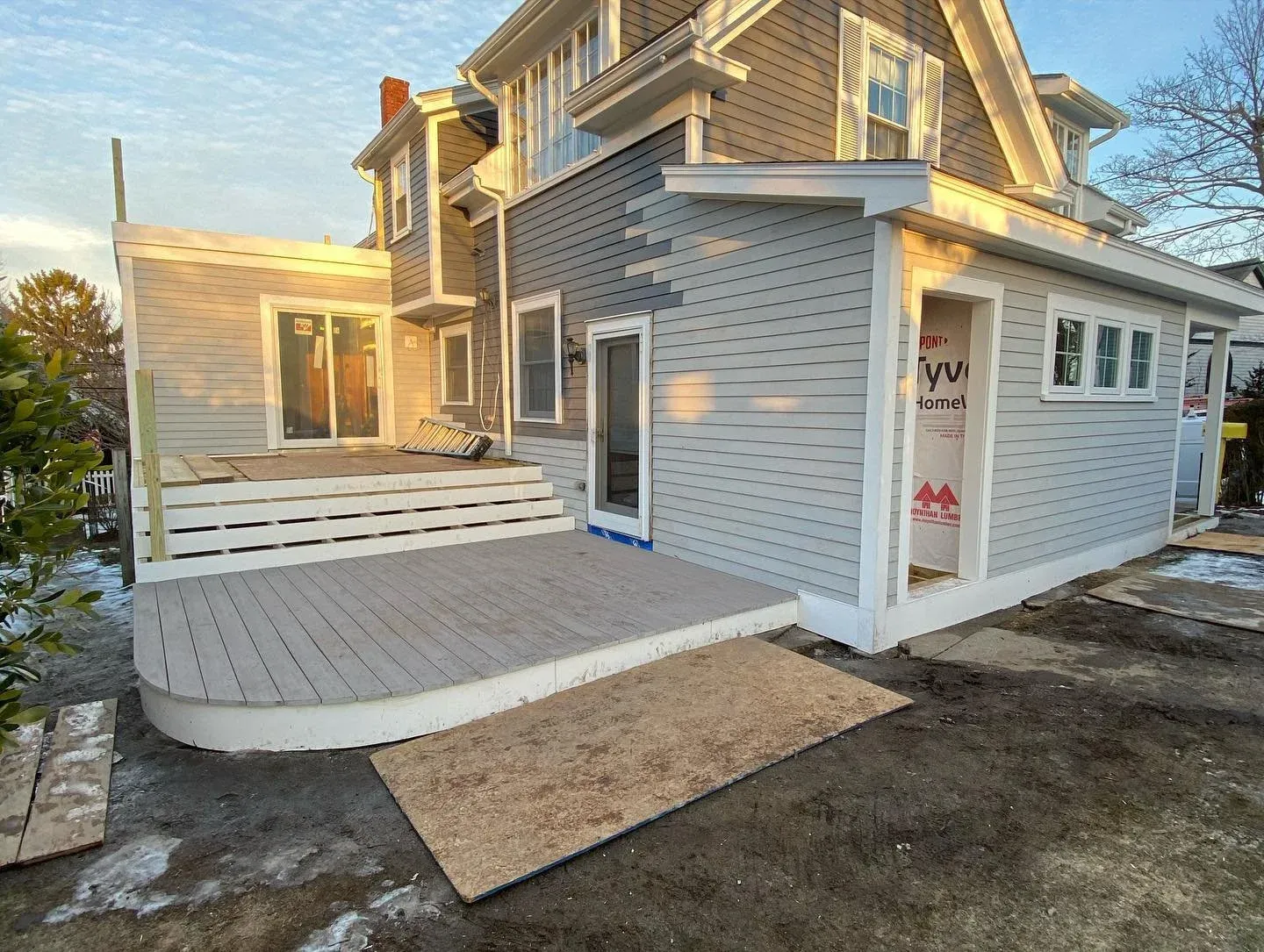 A house under construction with a wooden deck and sliding glass door, unfinished doorway with house wrap, and partly completed gray siding exterior, with patches of snow on the ground.