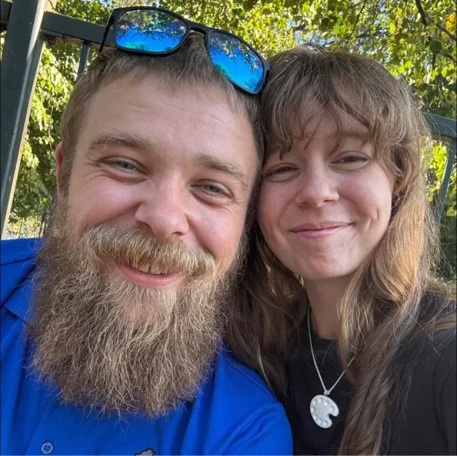 Close-up of a smiling young man with a beard wearing sunglasses on his head and a young woman with long hair and a necklace, outdoors with green trees in the background. These are the owners of Precision Radon Systems, a husband and wife team.