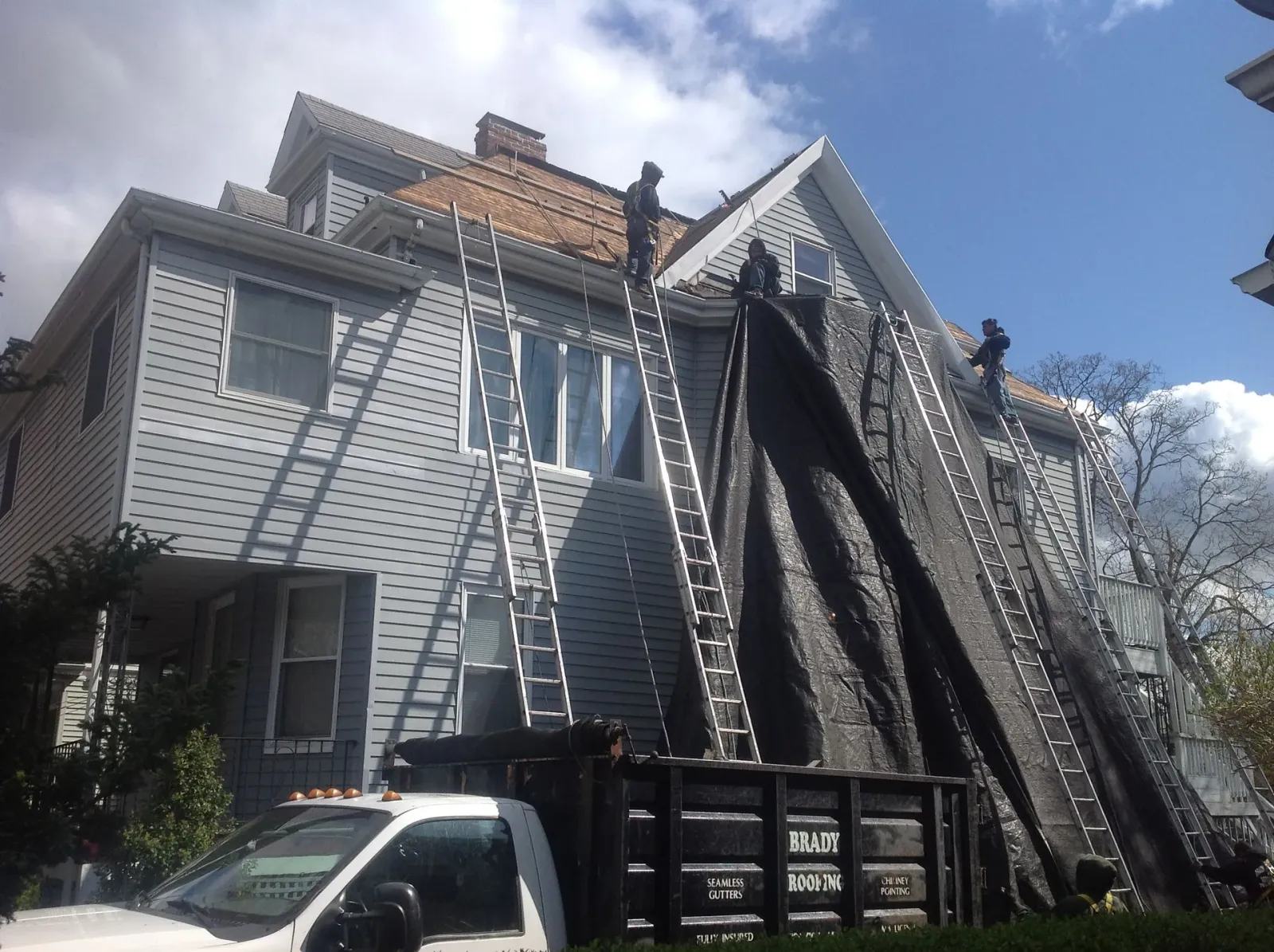 Laddered house with roof repair, large dumpster, and a work truck in front under a blue sky.