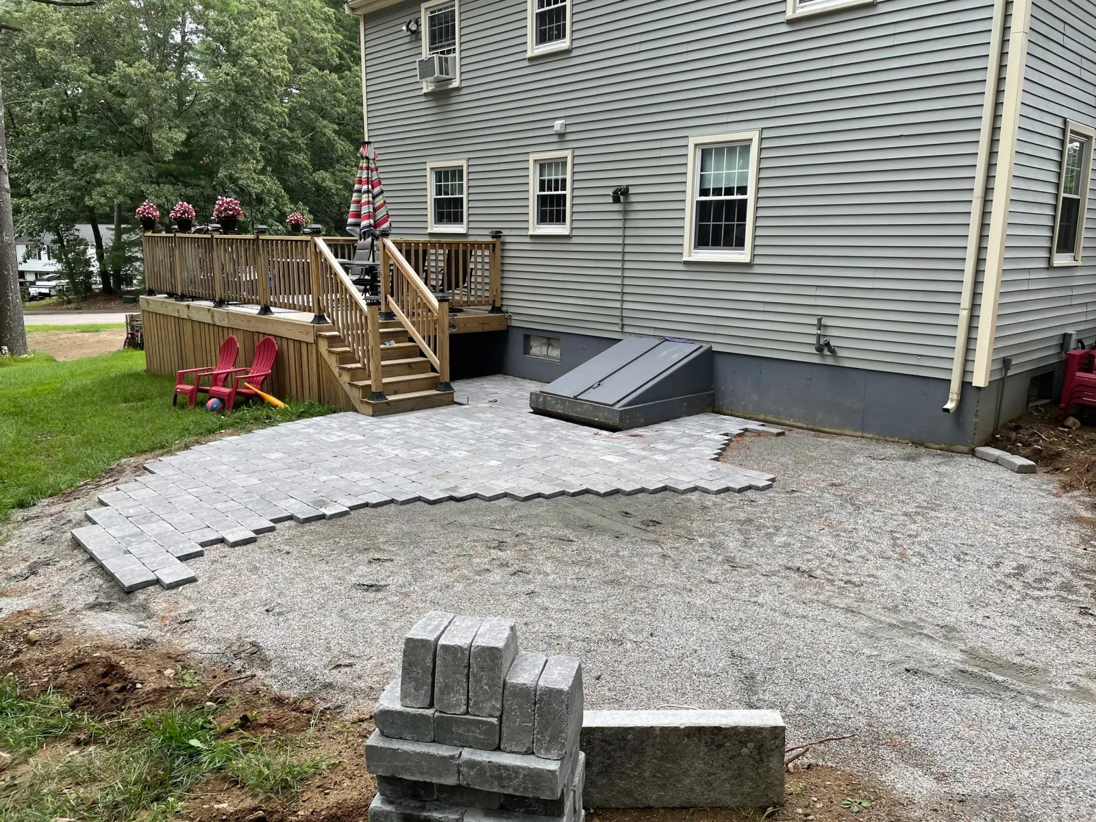 Backyard scene with a wooden deck, pink flowers, gray siding, under-construction patio with paving stones, and lawn chairs.