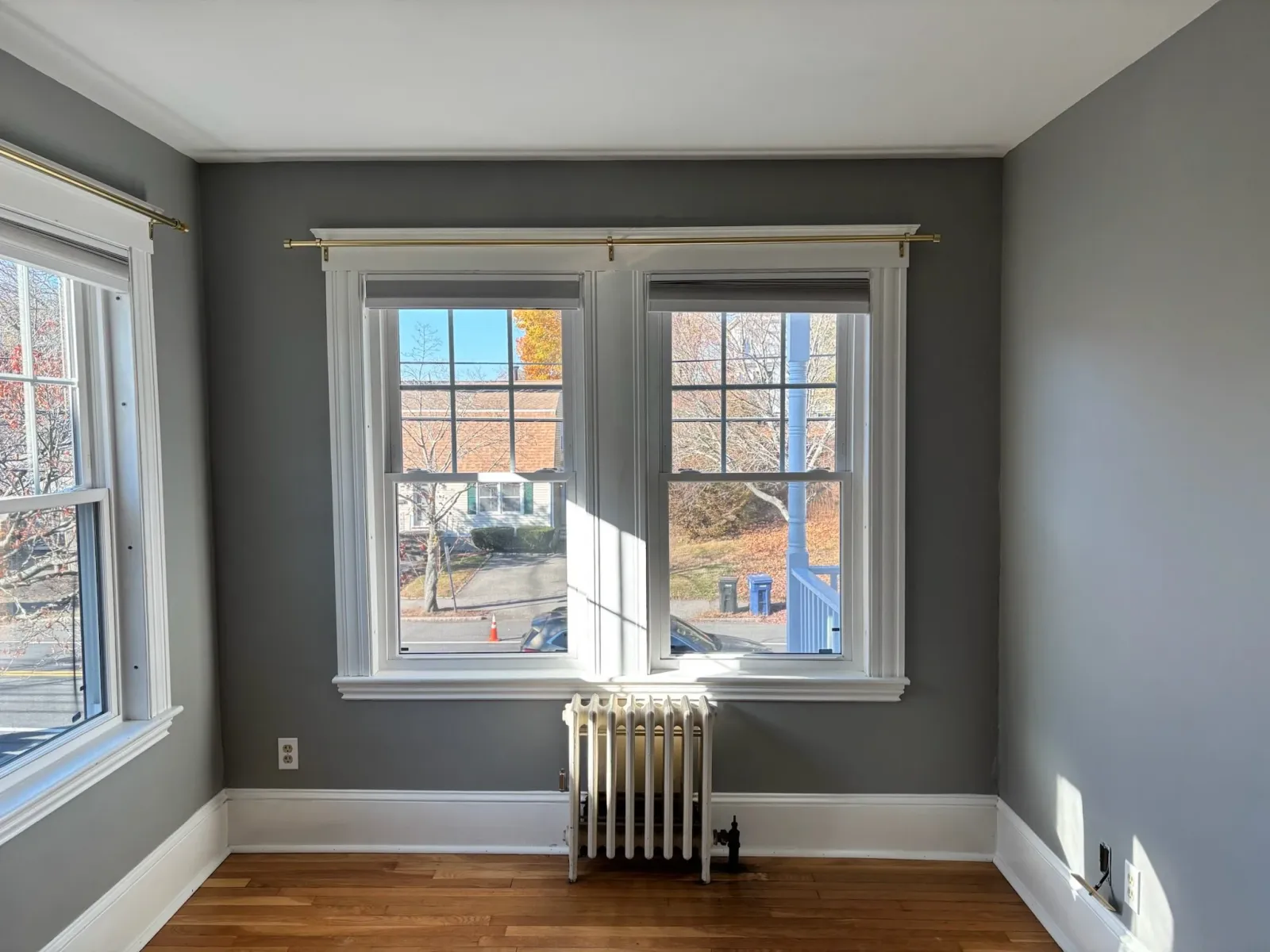 Empty room with gray walls, two large windows with white trim and curtain rods, a white radiator, and hardwood floors. Outside, there are trees and a blue recycling bin visible.