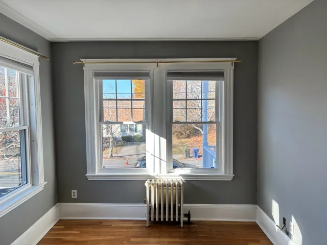 Empty room with gray walls, two large windows with white trim and curtain rods, a white radiator, and hardwood floors. Outside, there are trees and a blue recycling bin visible.