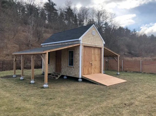 Small barn-like structure under construction with a ramp, surrounded by a grassy yard and a wooden fence, with a wooded hillside in the background.