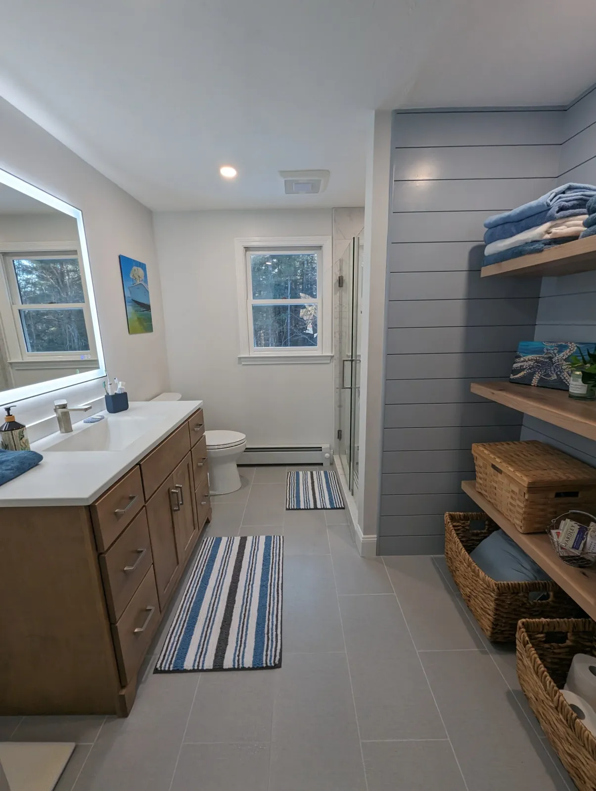 A modern bathroom with a wooden vanity, large mirror with built-in lighting, and gray tiled floor. There is a window with a view of trees, a small striped rug in front of the sink, a toilet, and a corner shower with glass door. A blue striped rug is on the floor, and built-in shelves hold towels and decorative items.