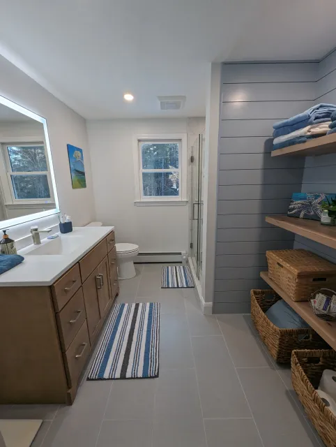 A modern bathroom with a wooden vanity, large mirror with built-in lighting, and gray tiled floor. There is a window with a view of trees, a small striped rug in front of the sink, a toilet, and a corner shower with glass door. A blue striped rug is on the floor, and built-in shelves hold towels and decorative items.