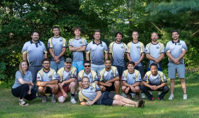 Team photo of a soccer team in gray and yellow jerseys, posed on grass in front of trees.