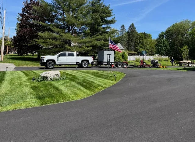 A white pickup truck parked on a paved driveway with a trailer attached, flying an American flag. In the background, several lawn equipment pieces, including mowers and other machinery, are lined up. There are large trees, a basketball hoop, a white fence, and a small garden with a rock and flowers, under a sunny sky.
