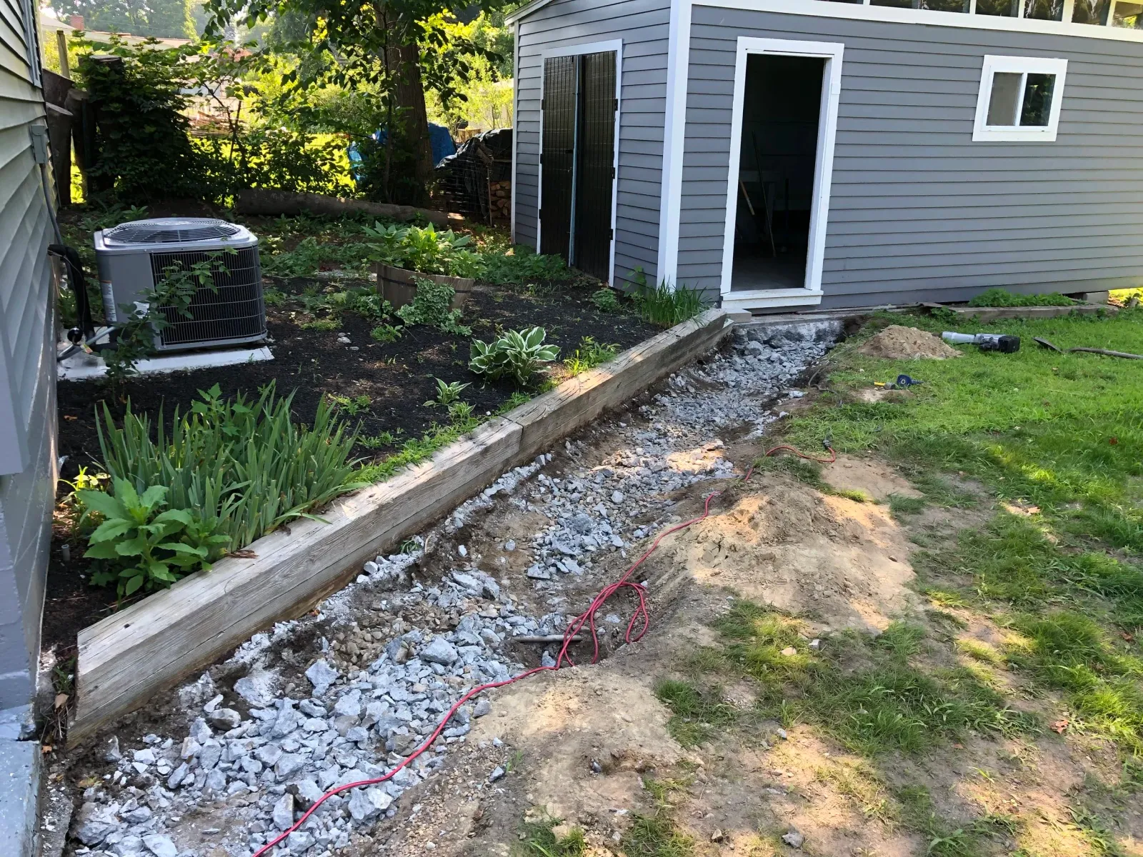 Backyard with construction site beside a gray shed; includes a gravel-filled trench, air conditioning unit, plants, and red electrical cord.