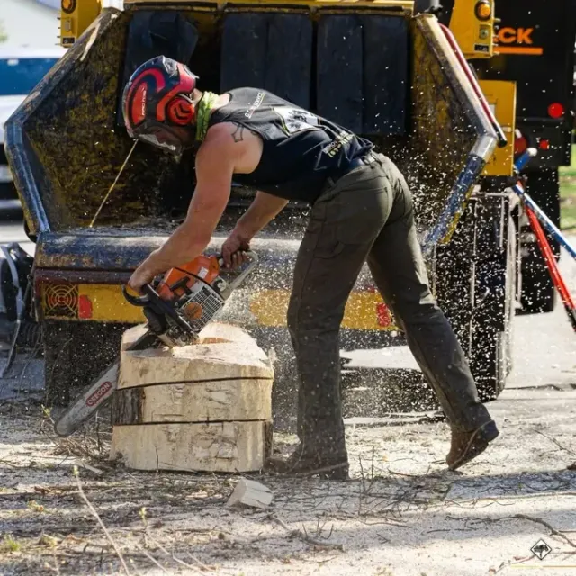 Person using a chainsaw to cut wood logs, wearing safety gear, near a wood chipper.