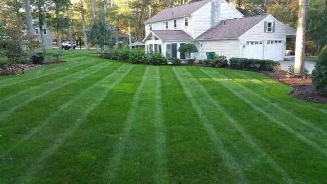 A two-story, light-colored house with a front porch and a neatly mowed, striped lawn featuring mulched landscaping beds.