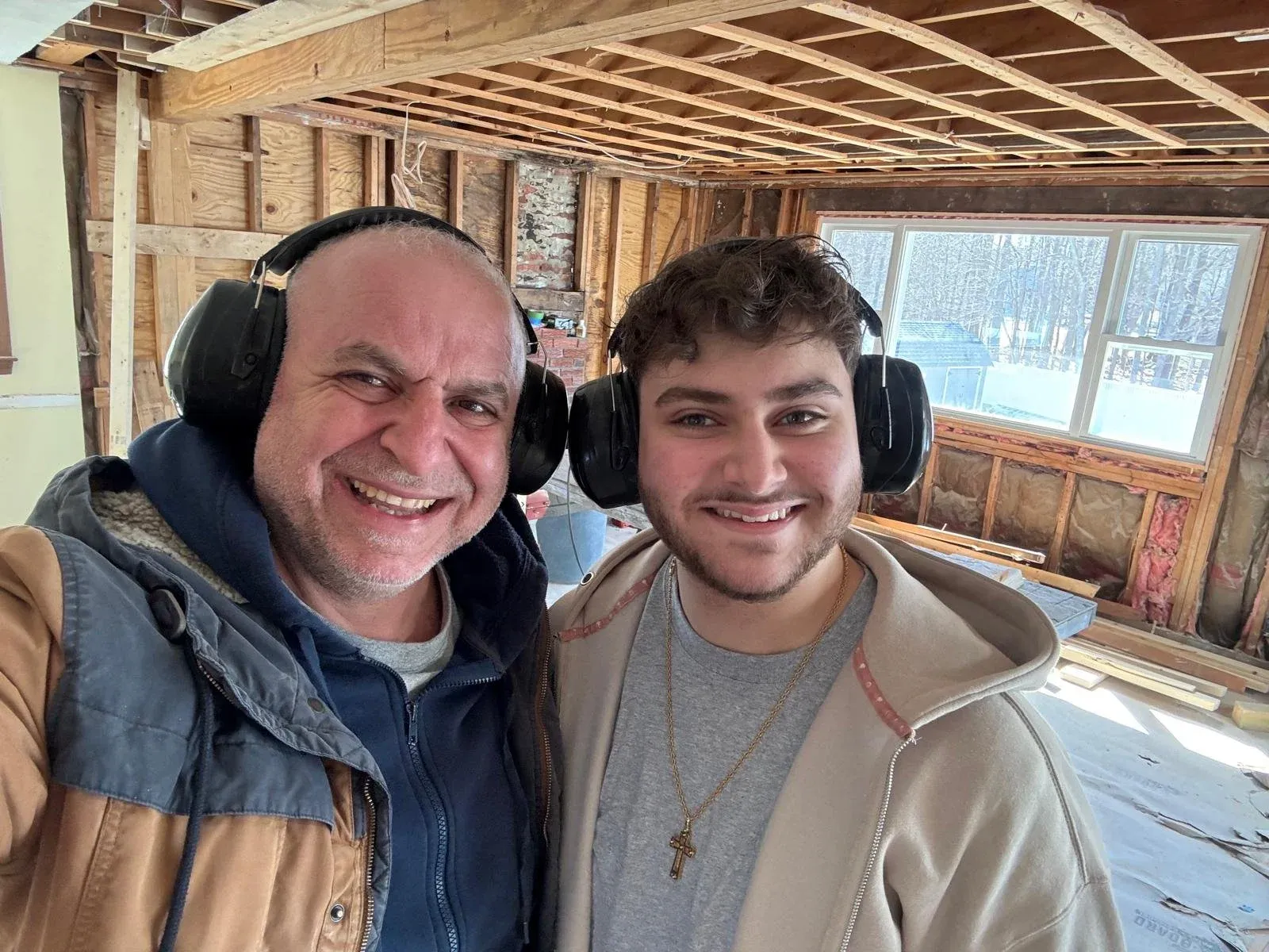 Two men wearing noise-canceling headphones smiling for a selfie inside a house under construction, with exposed wooden beams, insulation, and a large window showing a snowy outdoor scene.