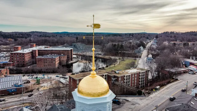 View of a town with a mix of historic and modern buildings, a pond, and a hilly landscape in the background, as seen from a high vantage point with a golden dome and weather vane in the foreground.