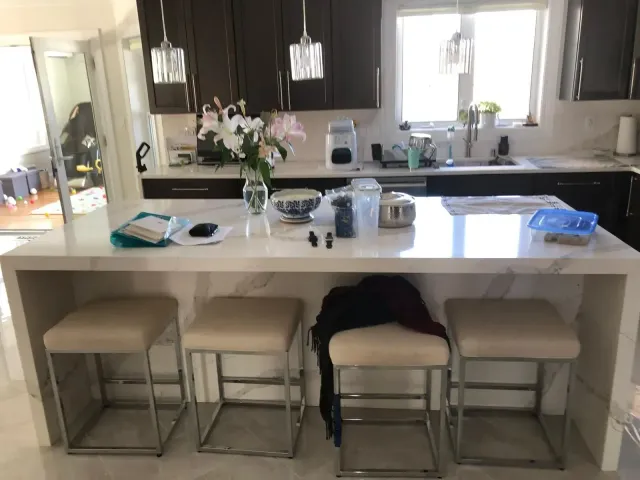 A kitchen with a white countertop island and four beige stools. Dark cabinets and a window are in the background.