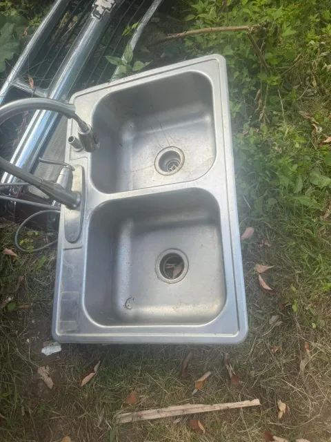 Outdoor stainless steel double-basin sink lying on grass beside hoses and a stick