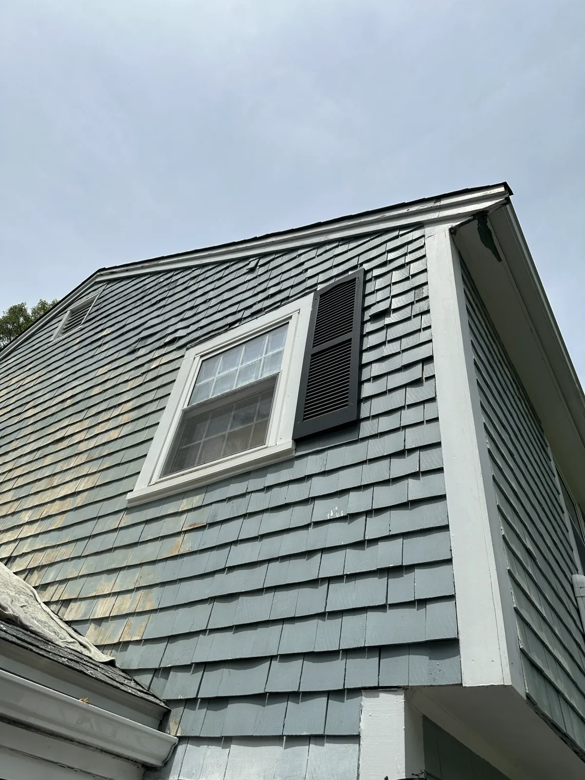 View of the upper corner of a house with gray shingle siding, white trim, a window with a white frame, and a black vented shutter, against a cloudy sky.