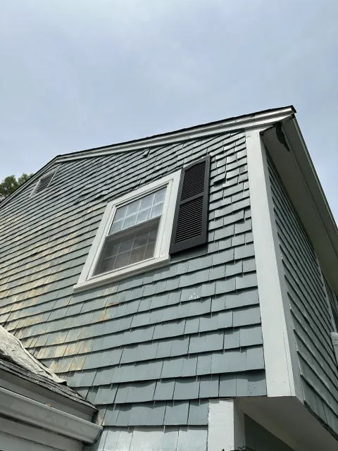View of the upper corner of a house with gray shingle siding, white trim, a window with a white frame, and a black vented shutter, against a cloudy sky.