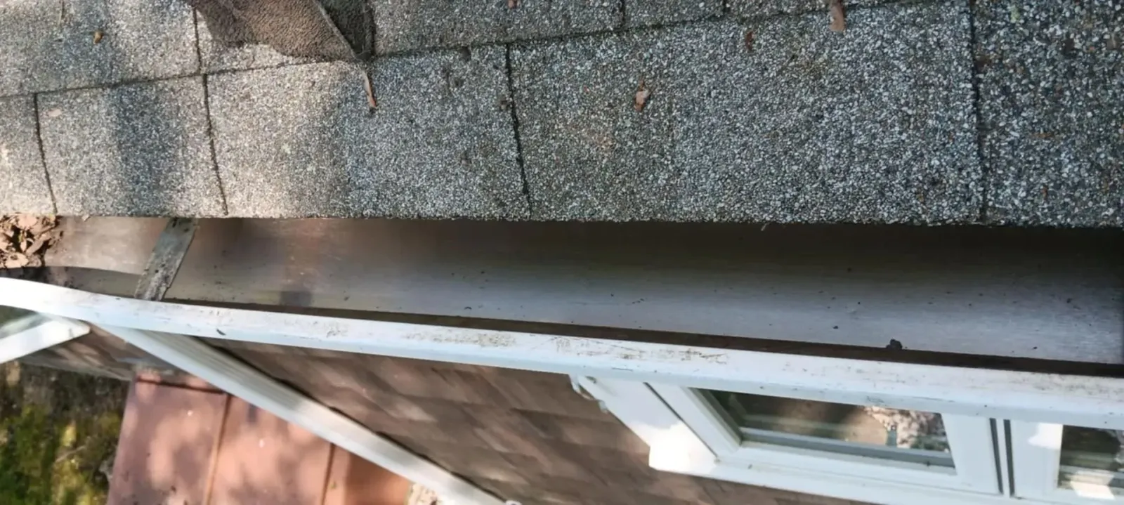 Roof shingles beside a white house exterior and gutter, with trees reflected in a window