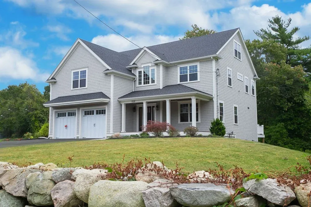 A large two-story house with gray siding, a dark shingle roof, and a attached garage, situated on a grassy lawn with a rock border.