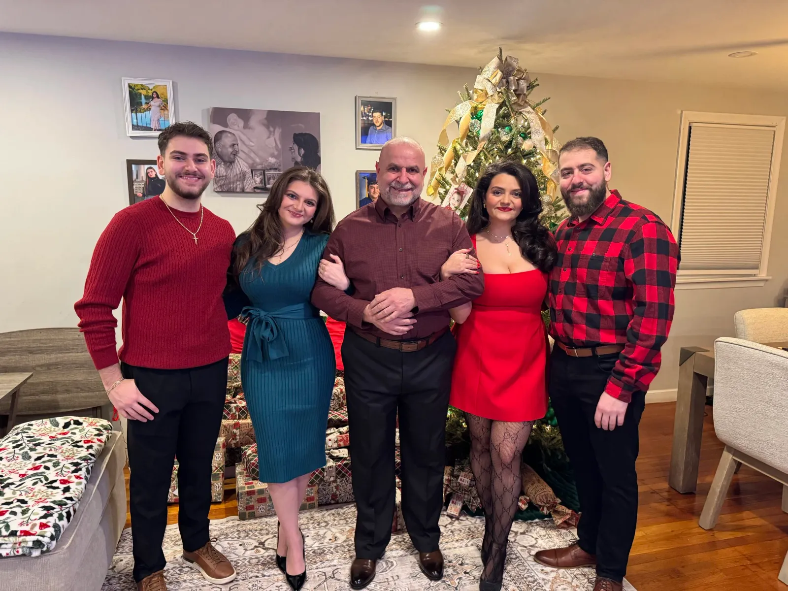 A family of five standing together in a living room decorated for Christmas with a Christmas tree and wrapped presents. The family includes two men, two women, and a man with a mustache and beard. They are all dressed in holiday attire, smiling, with the Christmas tree in the background.