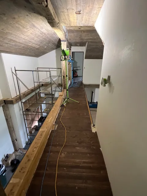 Interior of a house under renovation, with a narrow wooden hallway, construction tools, and scaffolding in the space.
