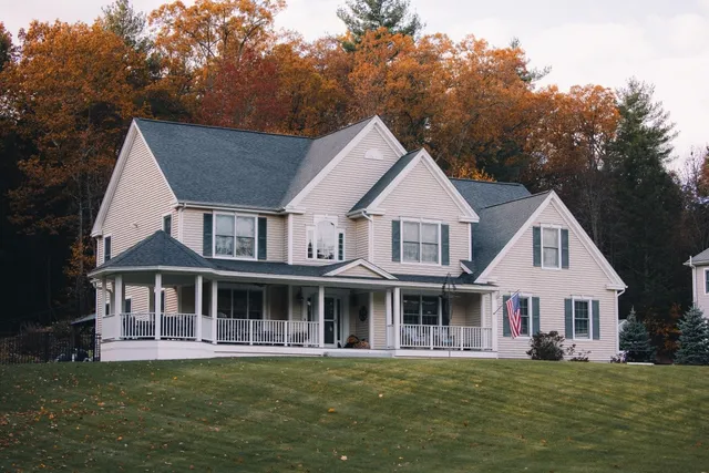 Two-story beige house with a front porch, dark roof installed by a reputable roofing company, American flag, and lawn, surrounded by autumn trees with orange foliage in the background.