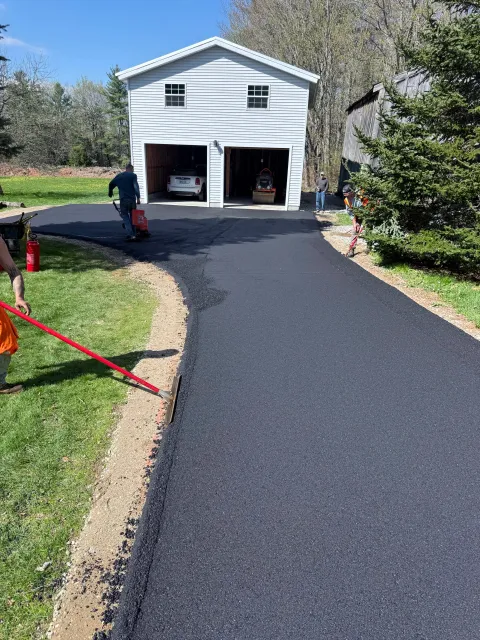 Workers paving a driveway in front of a white garage with two cars inside, surrounded by trees and clear blue sky.