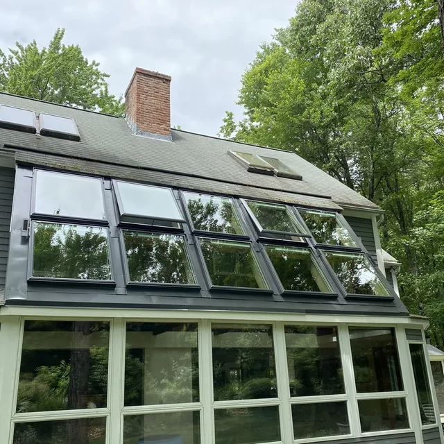 A sunroom exterior with a sloped glass roof featuring multiple open skylight windows against a wooded background.
