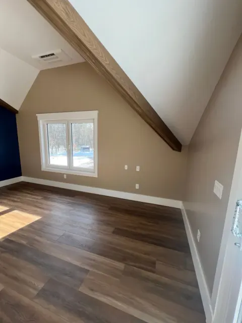 An empty room with tan walls, a slanted ceiling with a wooden beam, dark wood floors, and a window overlooking snow.